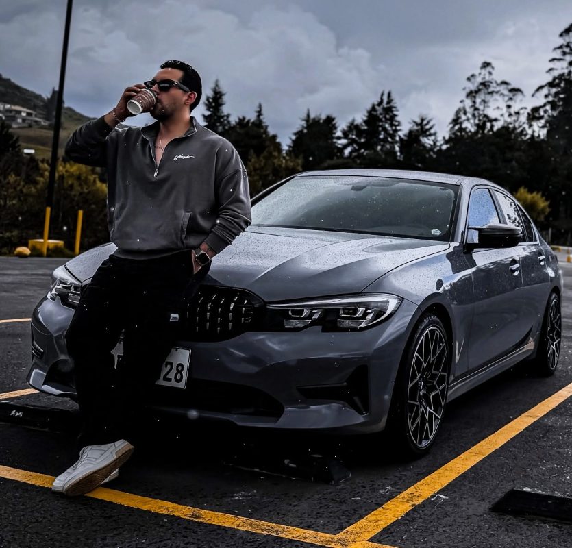 Hombre con gafas tomando café junto a un coche deportivo gris oscuro en un estacionamiento.
