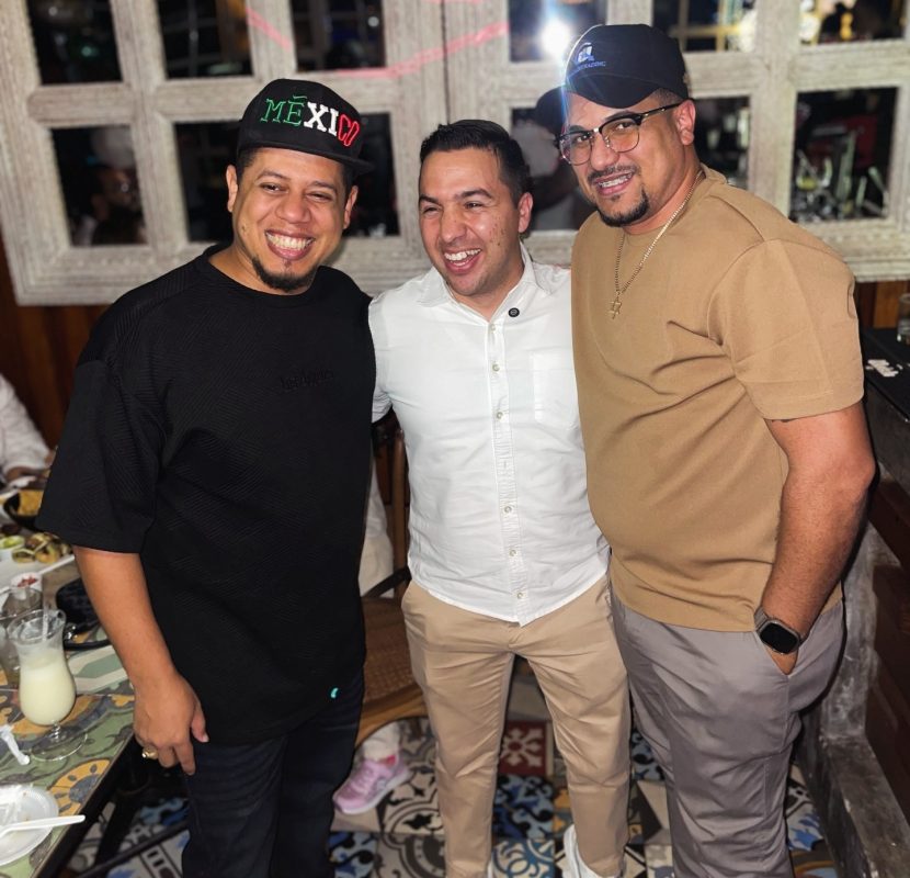 Tres hombres sonriendo en una reunión social, en un ambiente acogedor y decorado con espejos y mesas.